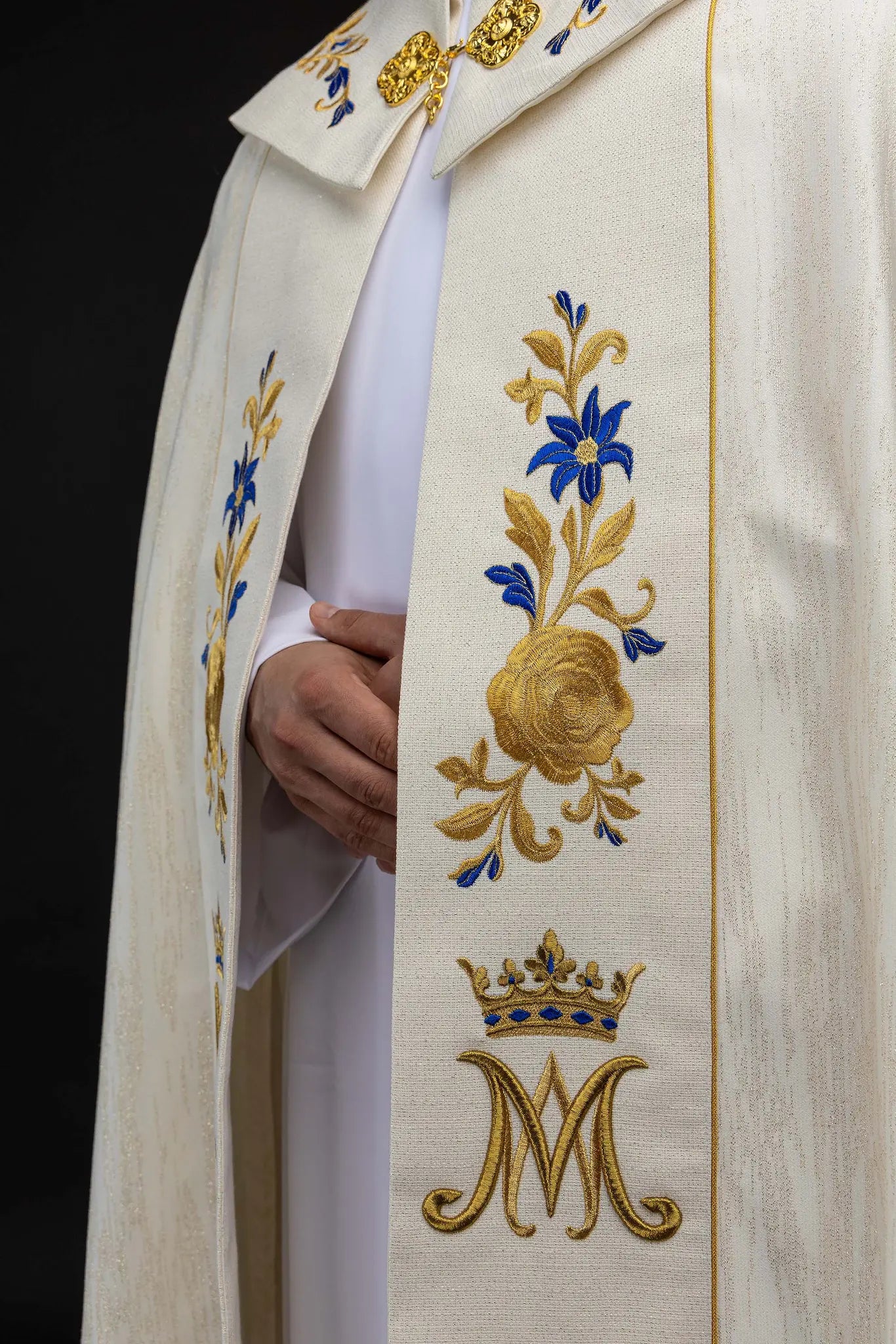 Ecru embroidered cope with Marian symbol, featuring gold and blue floral embroidery, a crowned 'M' monogram, and an ornate gold clasp with chain.