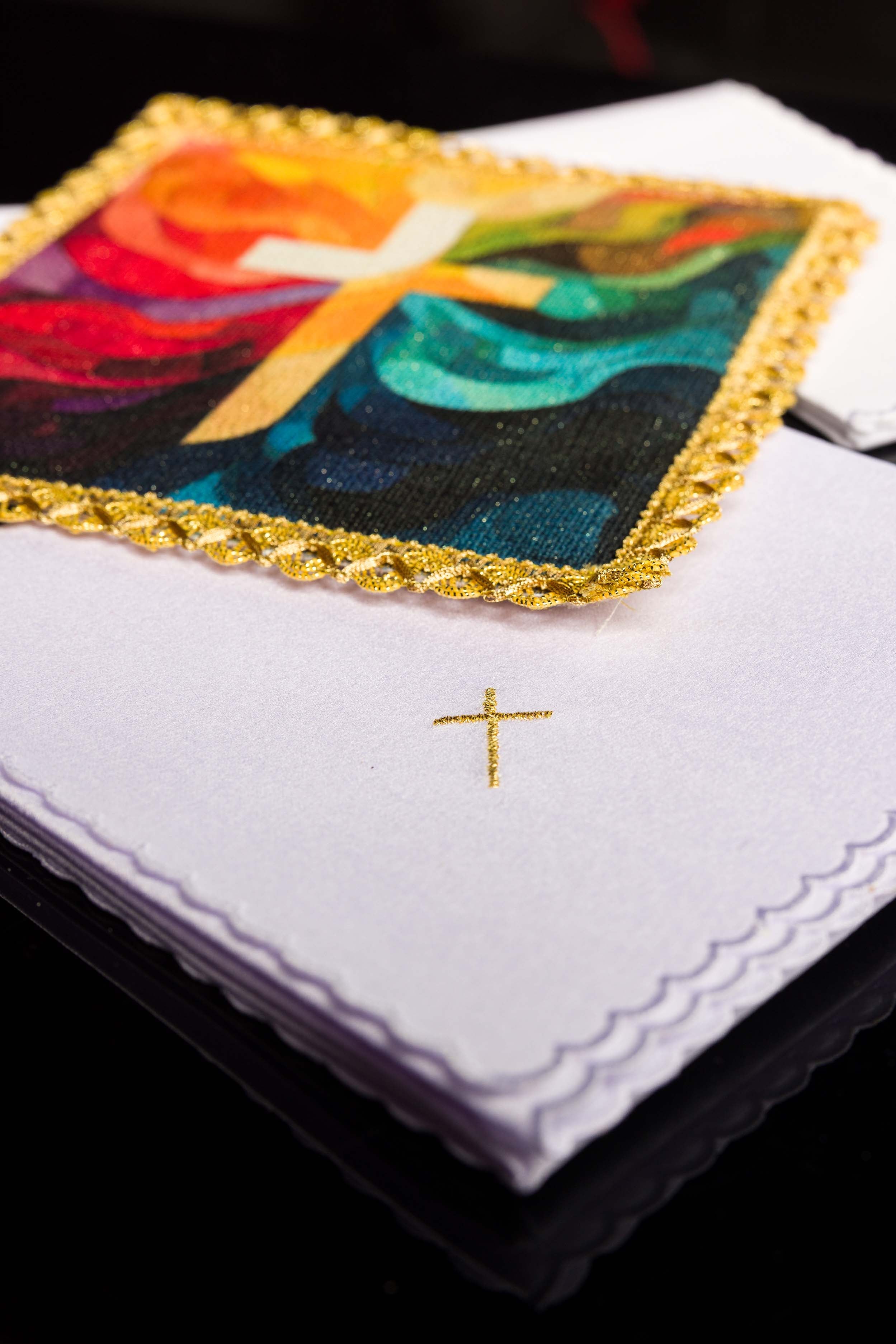 Folded white purificator with small embroidered gold cross next to the multicolour cross pall